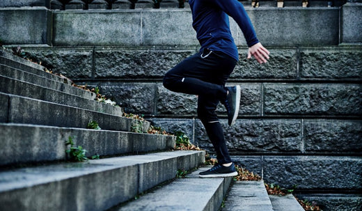 A runner in a blue long-sleeve shirt, black leggings, and black shoes is mid-step on worn outdoor stone stairs with leaves and grass. A stone wall in the background hints at an urban or park setting.