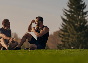 Two men, possibly father and son, are sitting on a grassy field. The younger man flexes his bicep while looking at the older man, both smiling.