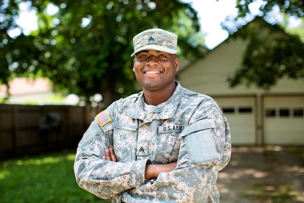 A smiling U.S. Army soldier in camouflage uniform and cap stands with arms crossed in front of a house and garage, with sunlight, trees, and a green lawn creating a warm, welcoming scene.