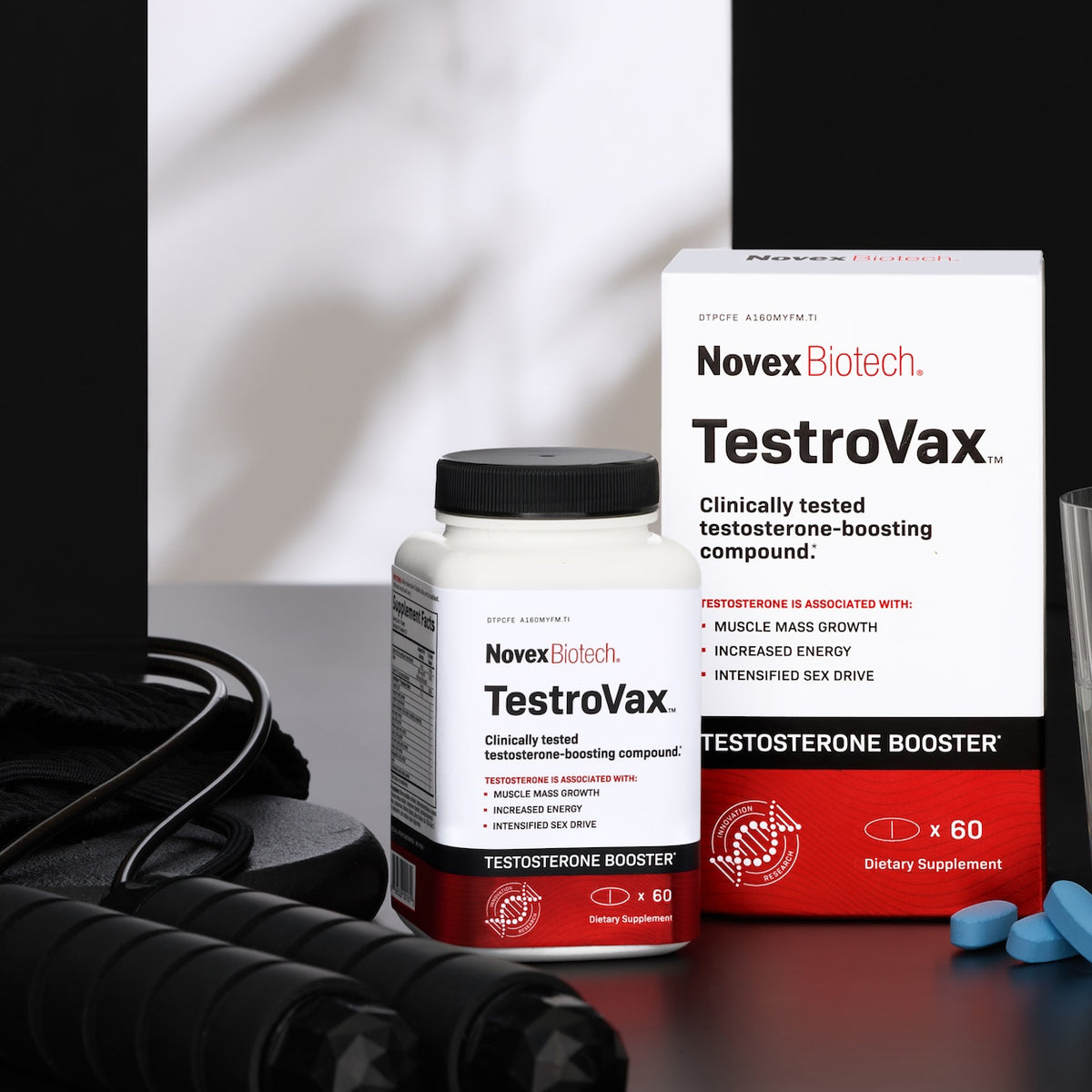 A bottle and box of Novex Biotech TestroVax testosterone booster are shown with a jump rope, pills, and workout gear on a black surface.