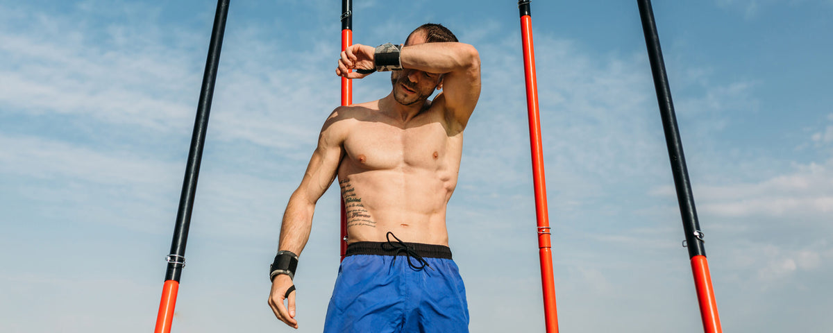 A muscular shirtless man in blue shorts and wrist wraps wipes sweat from his brow under a blue sky. He stands between orange and black poles, with a tattoo on his left arm, suggesting outdoor training.