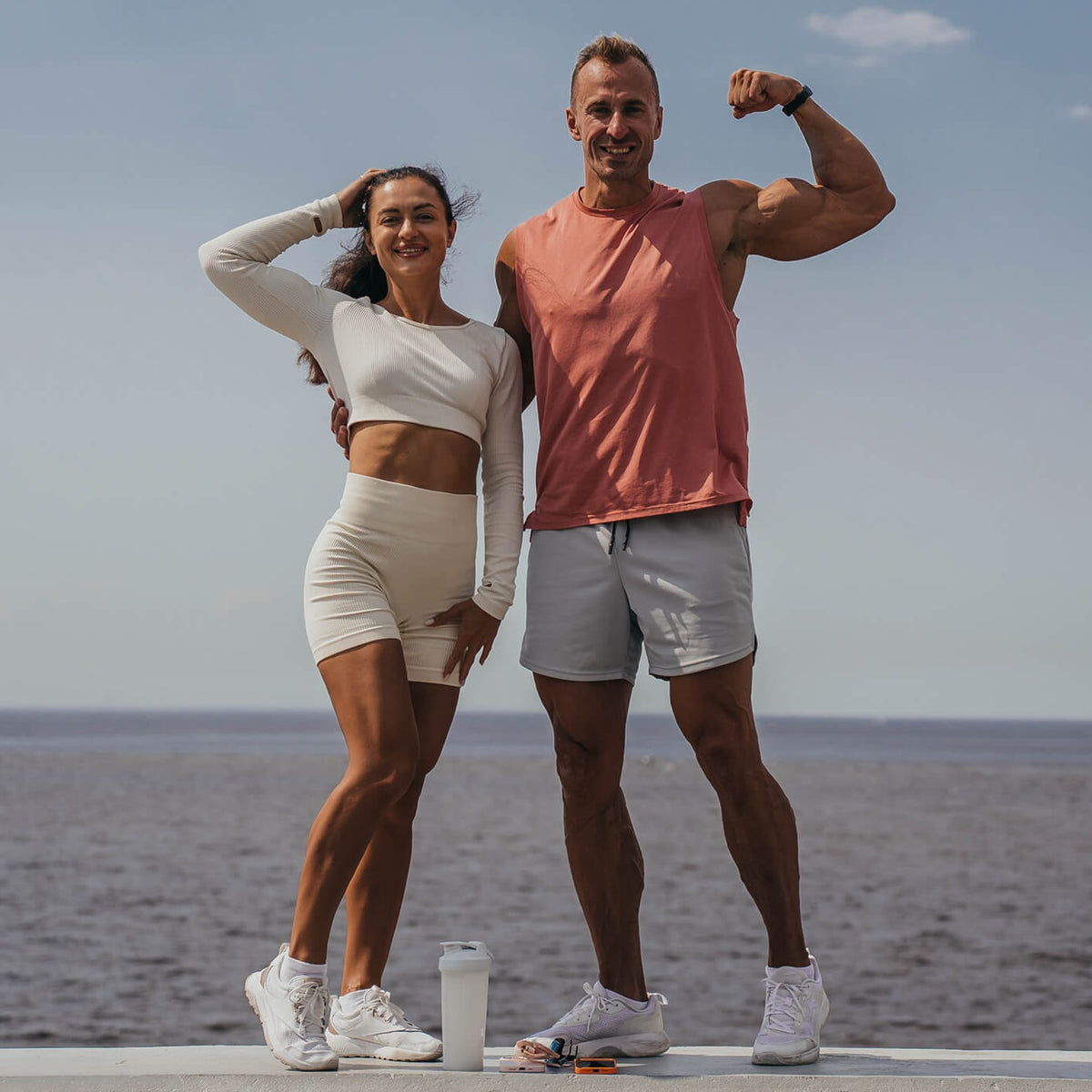 Two fit people pose by the water: a woman in a white crop top and shorts, and a man in a peach tank top and gray shorts flexing his bicep.
