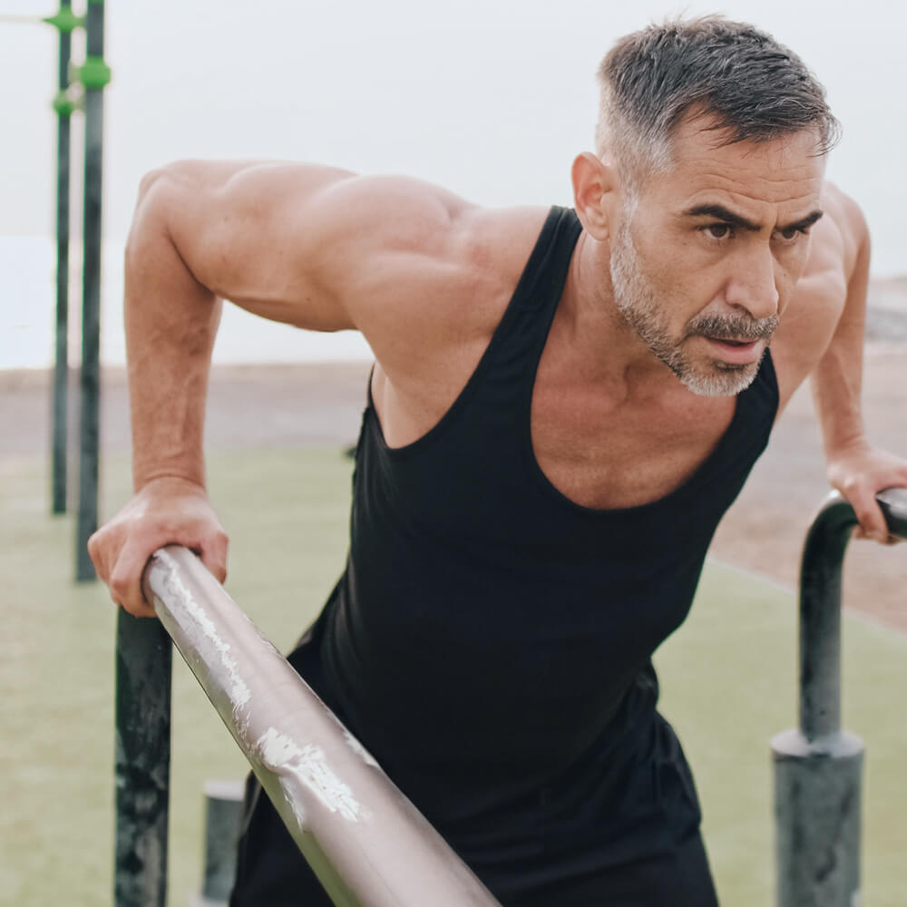 Man in a black tank top exercising outdoors on a metal bar.