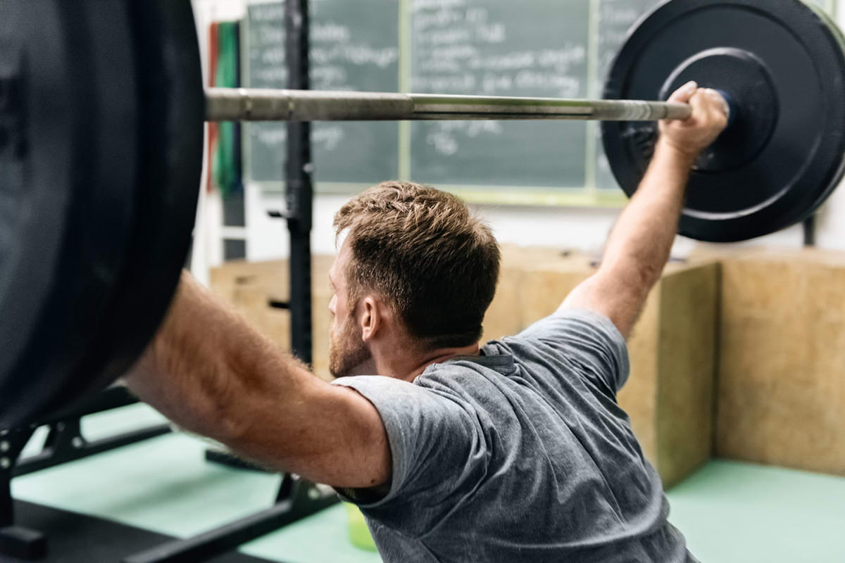 Fit white man with a beard in a gray t-shirt lifting barbell above his head surrounded by racks and gym equipment