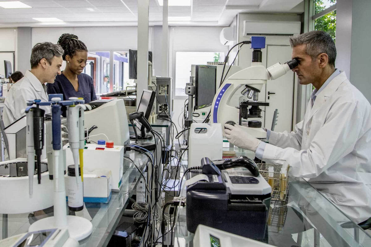 Medical research team in a modern laboratory, with a male scientist examining a sample under a microscope and two colleagues discussing data nearby