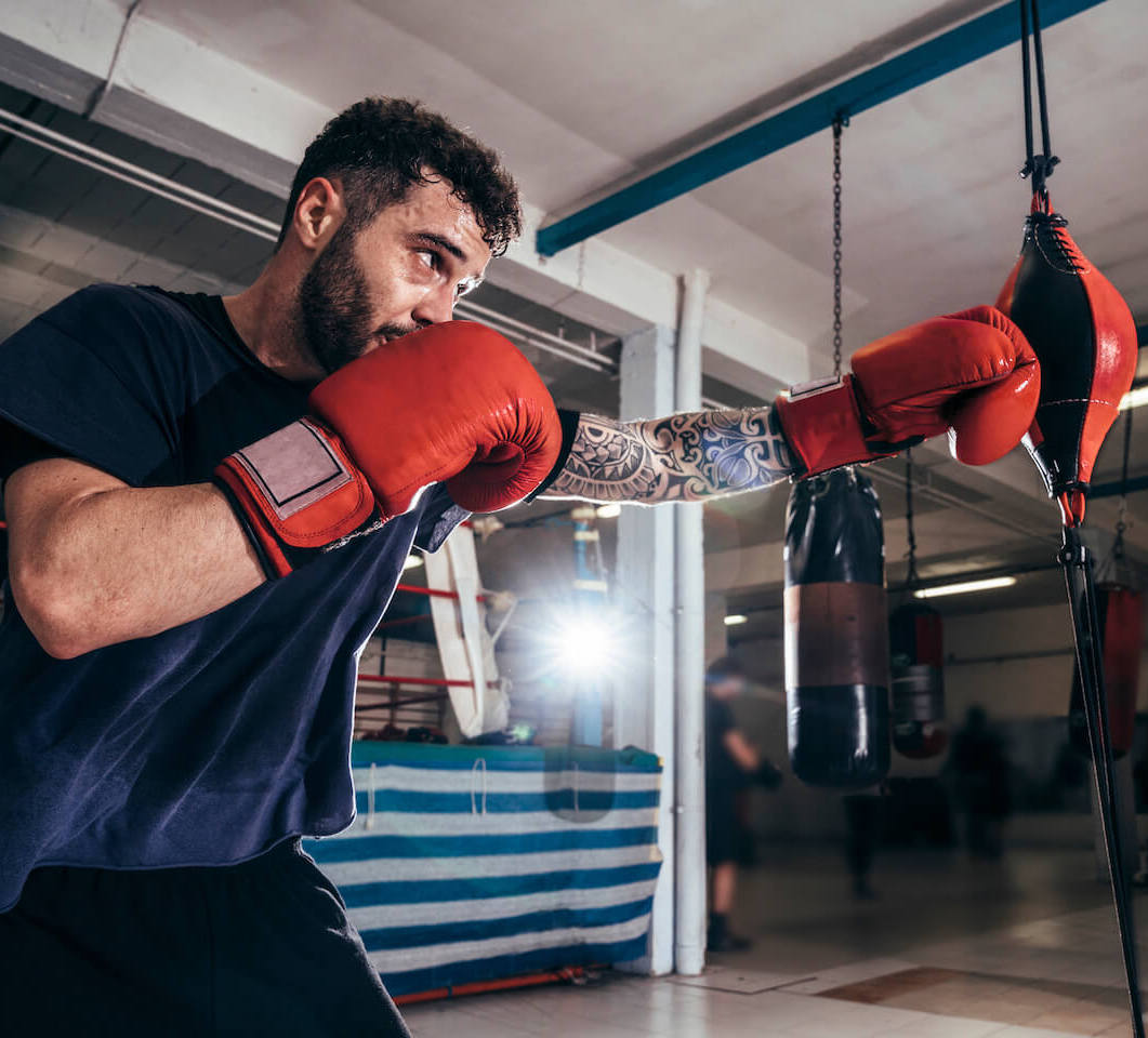 Man with a tattoo sleeve on one arm wearing boxing gloves hitting a speedbag