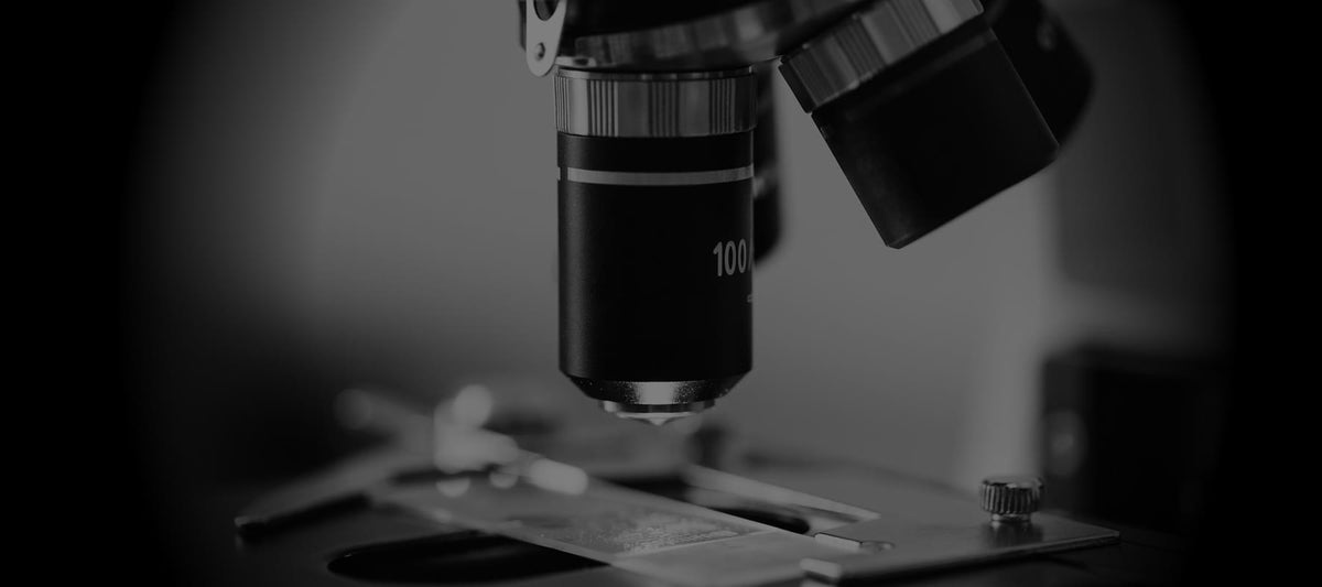 A black-and-white image of a microscope with a 100x magnification lens in focus, positioned over a slide on a stage. The background is blurred, emphasizing the microscope's objective lens and part of the slide holder, suggesting a laboratory or scientific research setting.