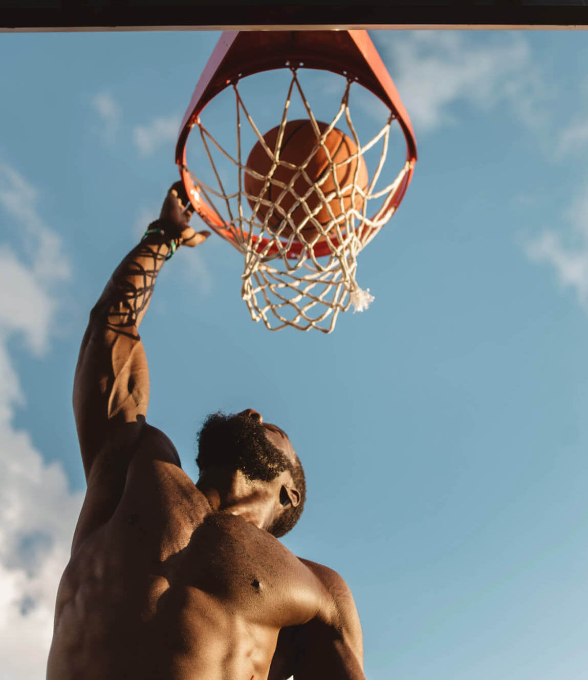 A muscular man performing a slam dunk on a basketball hoop, with the ball going through the net. He is shirtless, wearing a wristband, and the scene is set against a bright blue sky with scattered clouds. The image captures the action from below, emphasizing the height and power of the dunk.