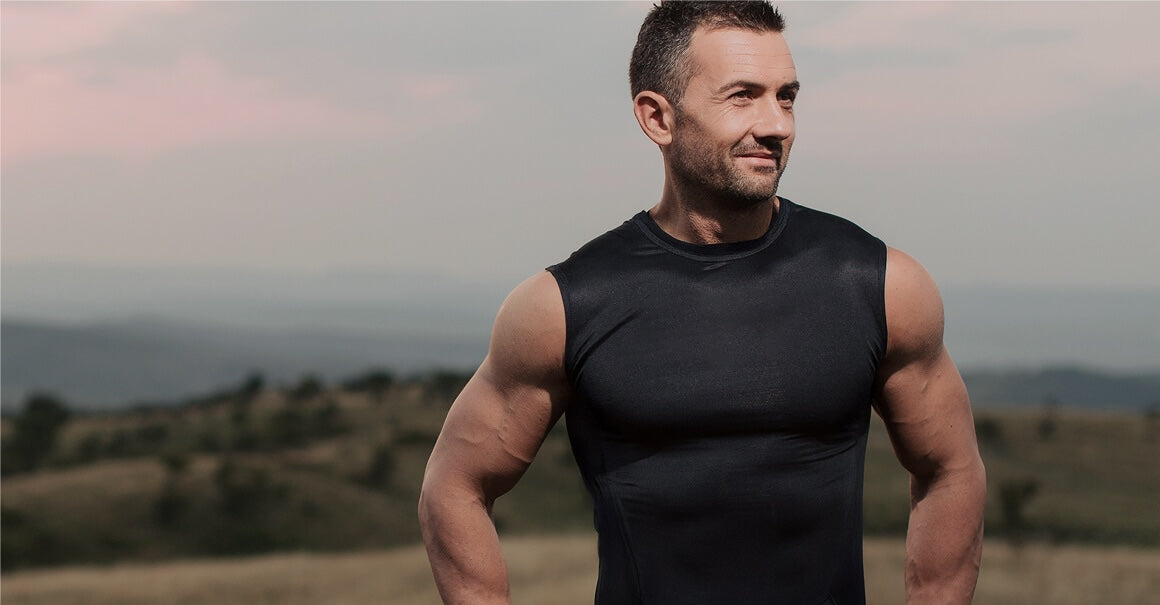 Muscular man in a black tank top, looking stoic, with a blurred outdoor background of mountains and hills.