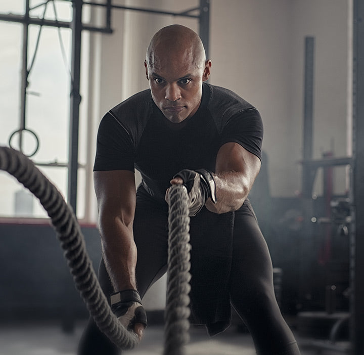 Athletic black man in gloves and athletic wear doing a battle ropes workout with gym equipment in the background
